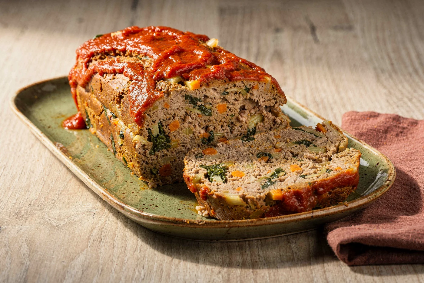 Asado alemán, un pan de carne cubierto con salsa de tomate y cortado en rodajas que muestran trocitos de acelga, zanahoria y apio, sobre una fuente alargada verde.