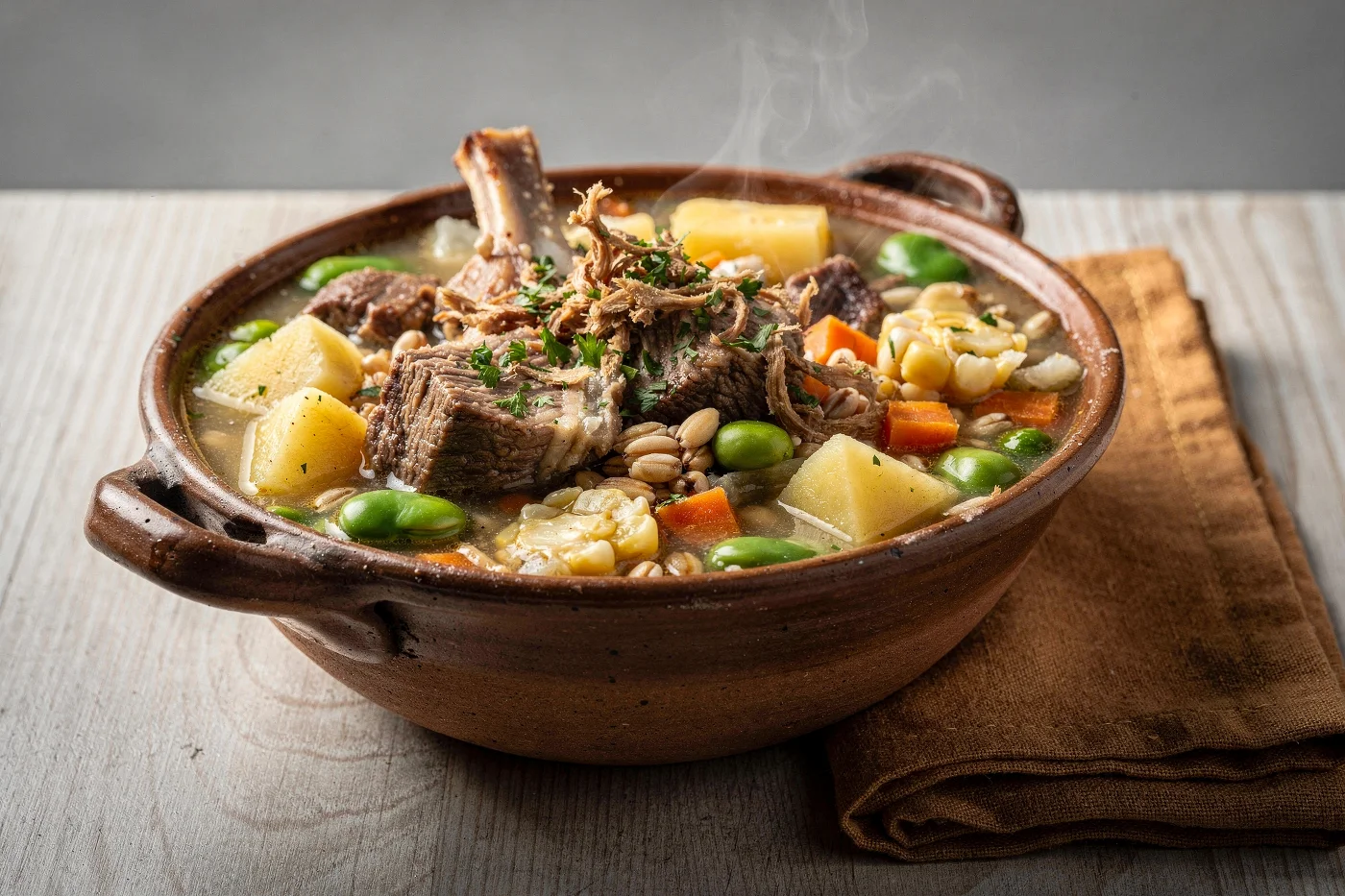 Cazuela de greda con chairo andino: trozos de carne con hueso y carne deshilachada, papa, habas, zanahoria, maíz y mote en un caldo con perejil picado, sobre una mesa de madera clara con un paño café.