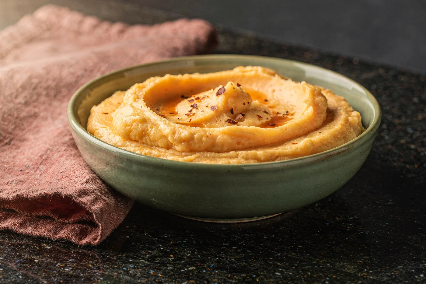 Cuenco verde con puré picante cremoso, con un poco de mantequilla derretida y ají seco en hojuelas encima, sobre una encimera oscura con un paño rosado.