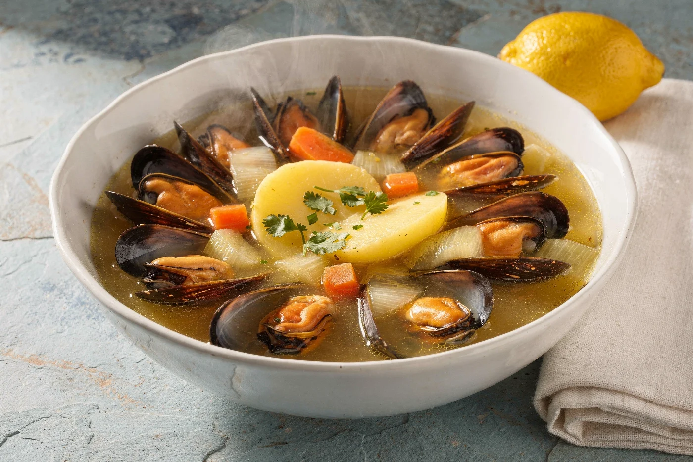 Plato hondo blanco con sopa de choritos: choritos abiertos en su concha alrededor de una rodaja de papa con perejil, con zanahoria en cubitos y cebolla en el caldo, y un limón entero al lado.